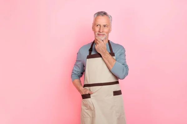 Fénykép a bájos pozitív nyugdíjas férfi viselni szürke ing kötény kéz kar áll keres üres tér elszigetelt rózsaszín háttér — Stock Fotó
