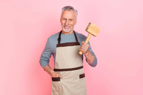Fénykép édes vicces nyugdíjas férfi viselni szürke ing kötény gazdaság kalapács elszigetelt rózsaszín szín háttér — Stock Fotó