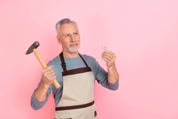 Foto de lindo hombre retirado de ensueño usar delantal de camisa gris sosteniendo martillo buscando espacio vacío aislado color rosa fondo — Foto de Stock