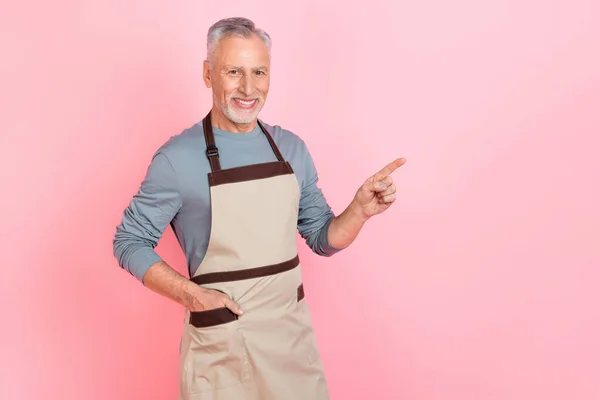 Fénykép a vonzó, csinos nyugdíjas férfi viselni szürke ing kötény mutató üres tér elszigetelt rózsaszín háttér — Stock Fotó