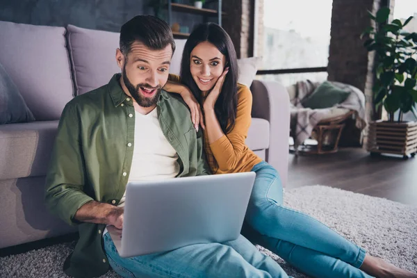 Retrato de hermosa pareja hermosa alegre asombrado sentado en la alfombra utilizando el ordenador portátil en casa plana en el interior —  Fotos de Stock