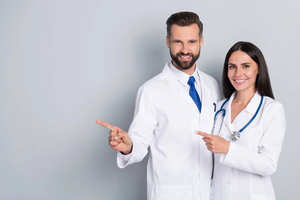 Foto de positivo bonito dos médicos usan abrigos blancos apuntando espacio vacío abrazando fondo de color gris aislado — Foto de Stock