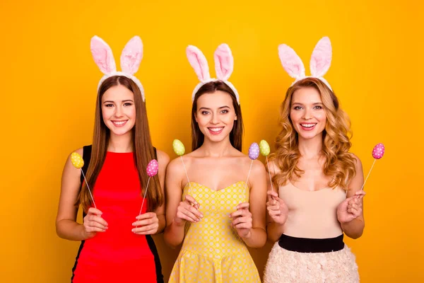 Foto rozkošné tři sestry dámy drží malované vejce hole dekor nosit načechrané králík uši pás chlad izolované žluté barvy pozadí — Stock fotografie