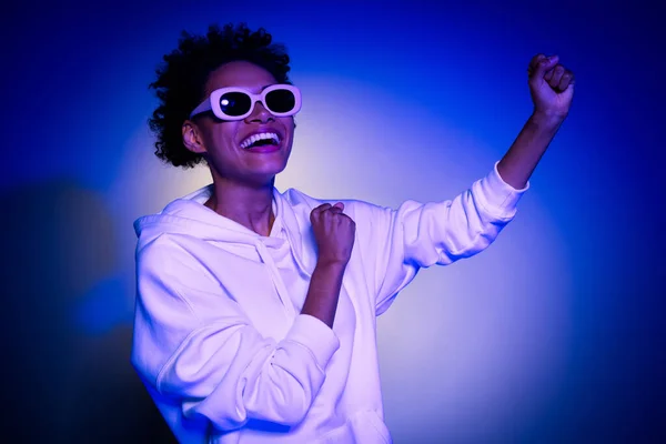 Foto di donna affascinante lucido indossare felpa occhiali scuri ballare avendo divertimento isolato sfondo di colore blu — Foto Stock