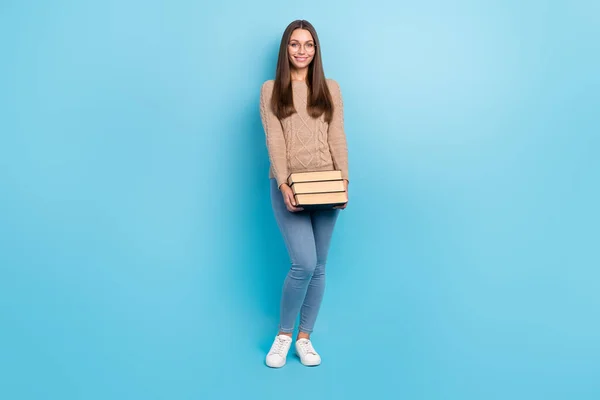 Tamanho do corpo de comprimento total vista de menina intelectual alegre atraente carregando livro pesado isolado sobre fundo de cor azul brilhante — Fotografia de Stock