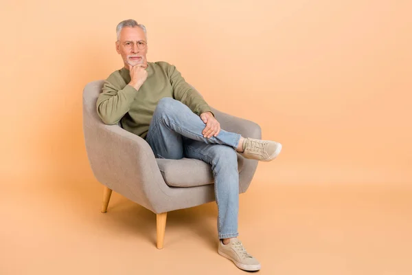 Foto de tamaño completo de estricto viejo gris peinado hombre sentarse desgaste gafas jersey jeans calzado aislado sobre fondo beige — Foto de Stock
