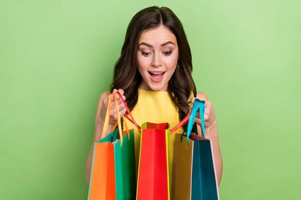 Ritratto di attraente ragazza allegra ragazza dai capelli ondulati guardando dentro borse nuove cose isolate su sfondo di colore verde brillante — Foto Stock