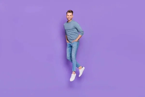 Фото привлекательного блестящего парня, одетого в серую рубашку, прыгающего высоко ходячие карманы рук изолированный фиолетовый цвет фона — стоковое фото