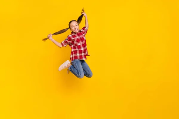 Foto de la encantadora colegiala emocionada usar camisa a cuadros saltando de brazos altos colas de caballo espacio vacío aislado color amarillo fondo —  Fotos de Stock
