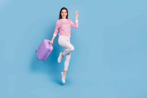 Full size portrait of attractive cheerful lady hold suitcase hand demonstrate v-sign isolated on blue color background — ストック写真