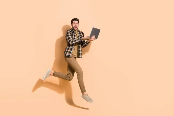 Full längd kroppsstorlek bild av attraktiv glad kille hoppa med hjälp av laptop kör isolerad på beige pastell färg bakgrund — Stockfoto
