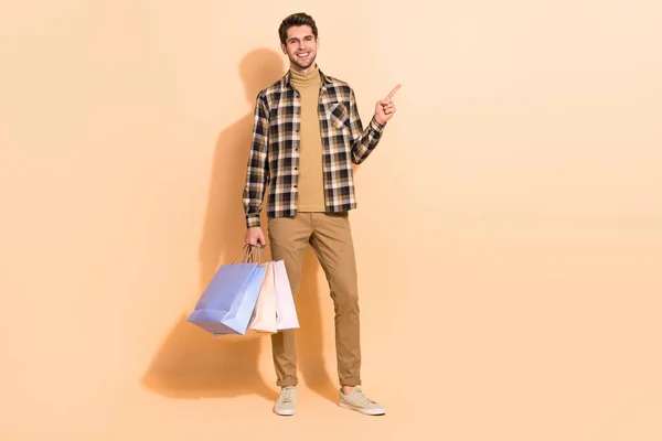 Vista completa del tamaño del cuerpo del atractivo chico alegre llevando bolsas demostrando oferta aislada sobre fondo de color pastel beige — Foto de Stock