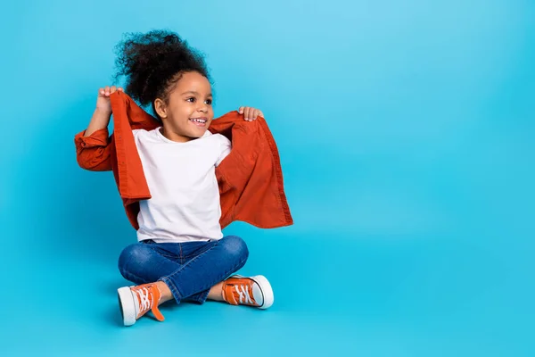 Foto de longitud completa de dulce ondulado niña pequeña sentarse look promocional usar camisa jeans zapatillas aisladas en fondo de color azul —  Fotos de Stock