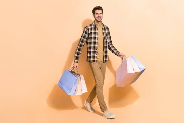 Вид в полный рост на привлекательного парня, заходящего в магазин в рубашке с сумками на бежевом фоне — стоковое фото