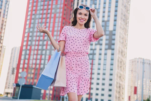 Fénykép a fiatal bájos nő viselni modern ruhát hold bevásárló táskák séta kéz érintés szemüveg szabadban — Stock Fotó