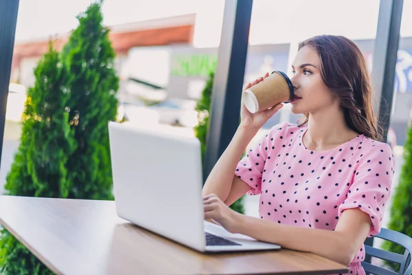 Porträt eines attraktiven welligen Mädchens mit Laptop, das aus der Ferne Latte trinkt und im Café im Freien sitzt — Stockfoto