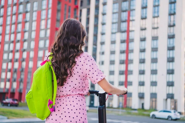 Traseira atrás vista de trás da menina de cabelos ondulados atraente vestindo mochila andar de scooter passar o fim de semana ao ar livre — Fotografia de Stock