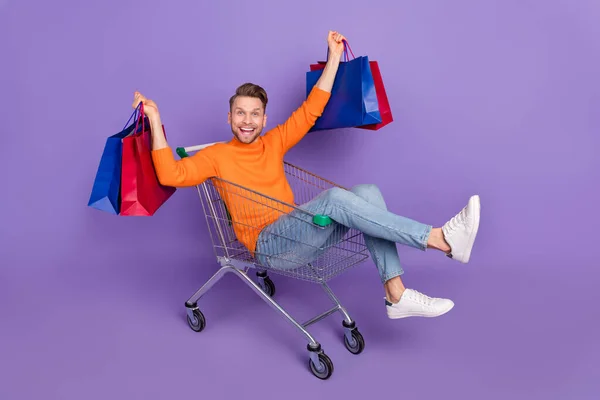Foto de cuerpo completo de alegre hombre atractivo lleno de alegría sentarse en carrito de compras a caballo aislado sobre fondo de color violeta — Foto de Stock