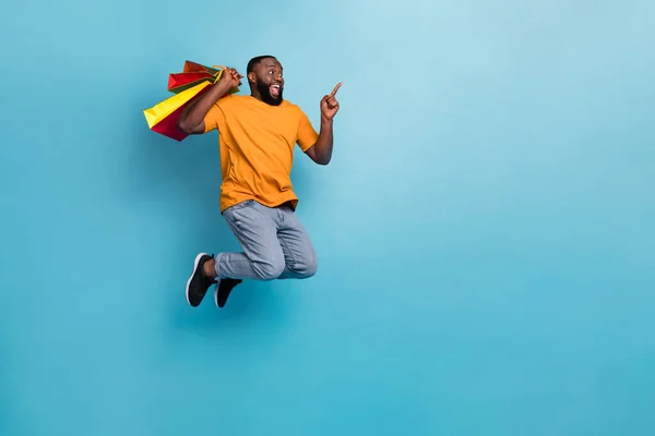 Photo of impressed funny guy dressed orange t-shirt jumping holding bargains pointing empty space isolated blue color background — ストック写真