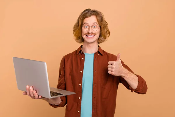 Retrato de chico guapo alegre de pelo rojo de moda utilizando el ordenador portátil que muestra el anuncio de pulgar aislado sobre fondo de color pastel beige —  Fotos de Stock