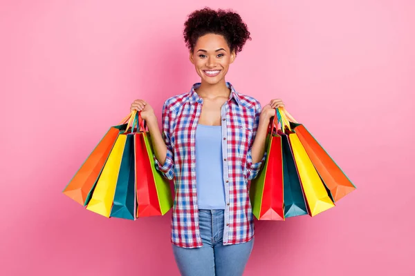 Foto de morena funky rizado peinado señora celebrar bolsas usar camisa a cuadros aislados en el fondo de color rosa — Foto de Stock