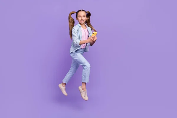 Foto de brilhante adorável menina da escola usar camisa jeans saltar digitando dispositivo moderno espaço vazio isolado cor roxa fundo — Fotografia de Stock