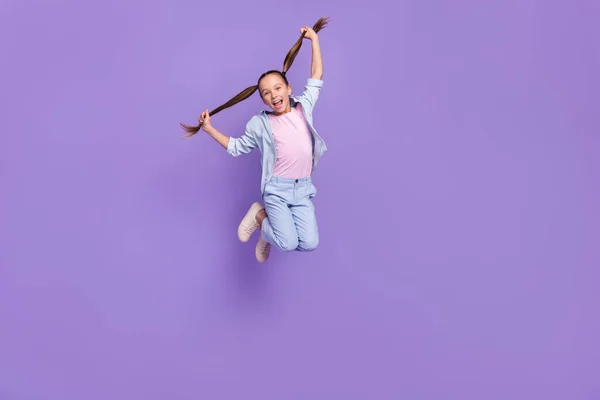 Foto de bastante alegre pequeña colegiala vestida jeans camisa sosteniendo colas saltando espacio vacío aislado color violeta fondo —  Fotos de Stock