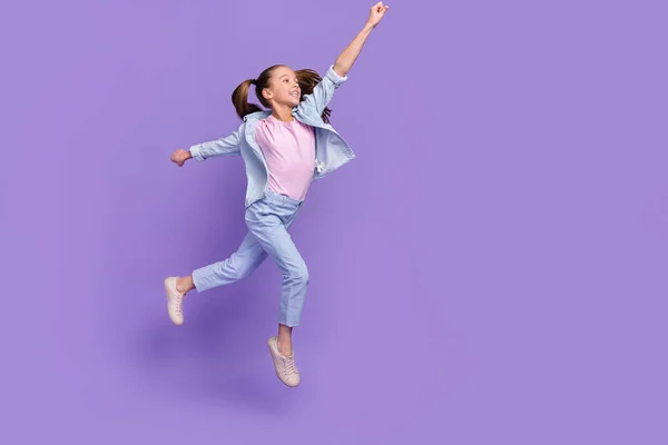 Фото милої збудженої маленької школярки в джинсовій сорочці, що стрибає високо тримає руку порожній простір ізольований фіолетовий кольоровий фон — стокове фото