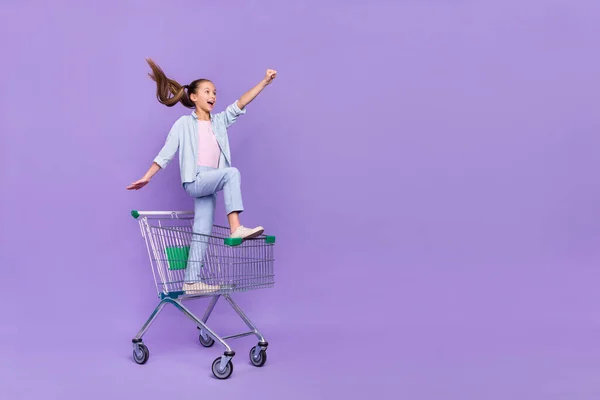 Fénykép vicces lenyűgözött kis iskoláslány öltözött farmer ing vezetés bevásárló tálca üres tér elszigetelt ibolya szín háttér — Stock Fotó
