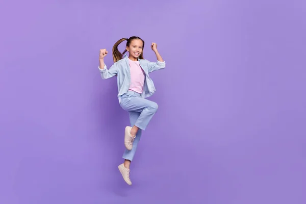 Foto de la niña de la escuela muy emocionada usar camisa de mezclilla puños en aumento saltando alto espacio vacío aislado color púrpura fondo —  Fotos de Stock