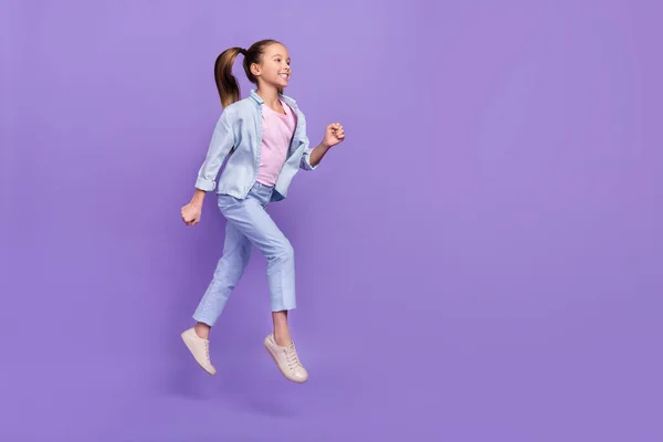 Foto de alegre doce pequena estudante vestida camisa jeans correndo rápido saltando espaço vazio isolado cor violeta fundo — Fotografia de Stock
