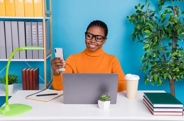 Foto von netten Fernarbeiter Dame sitzen Schreibtisch halten Telefon tragen Spezifikationen orange Pullover isoliert blaue Farbe Hintergrund — Stockfoto