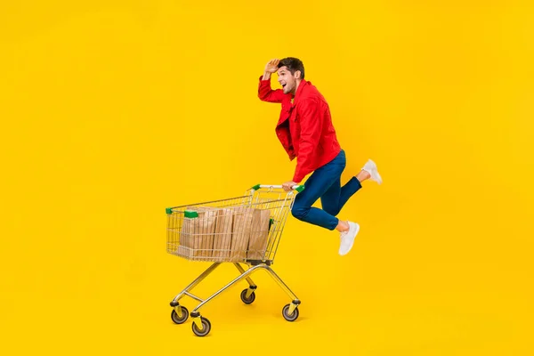 Full length body size view of attractive cheerful guy jumping carrying cart looking search goods isolated on bright yellow color background — ストック写真