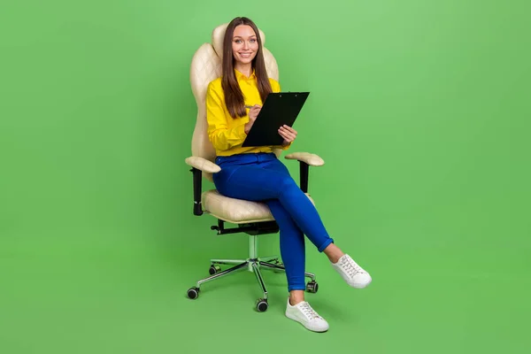 Vista completa del tamaño del cuerpo de la atractiva chica alegre sentada en la silla escribiendo lista de tareas aisladas sobre fondo de color pastel verde — Foto de Stock