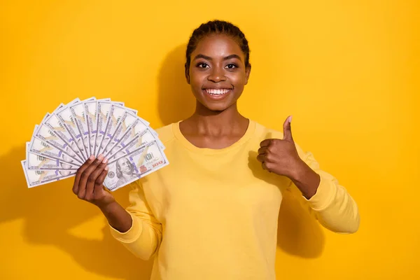 Portrait of beautiful trendy cheerful girl holding banknotes us atm cash showing thumbup isolated over bright yellow color background — Stock fotografie