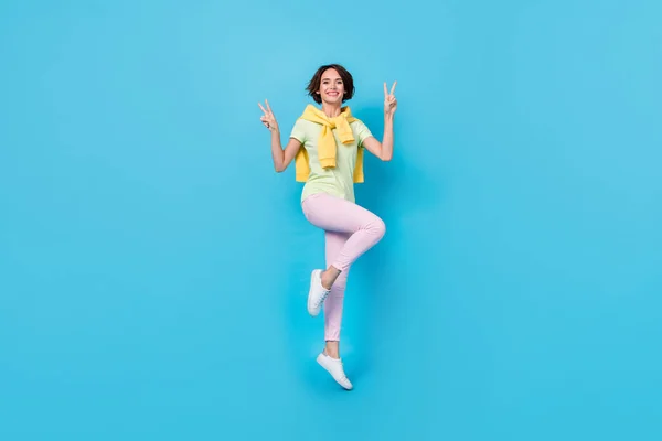 Full size photo of impressed millennial brunette lady jump show v-sign wear t-shirt trousers sneakers isolated on blue background — 스톡 사진