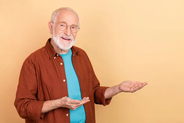 Foto de hombre de pelo blanco envejecido agradable celebrar espacio vacío usar gafas camisa marrón aislado sobre fondo de color beige — Foto de Stock