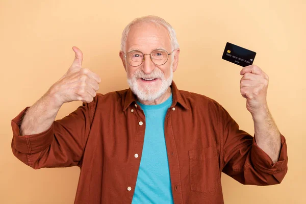 Photo of cheerful pensioner man showing thumb-up promote recommend plastic card isolated on beige color background — Stock Photo, Image