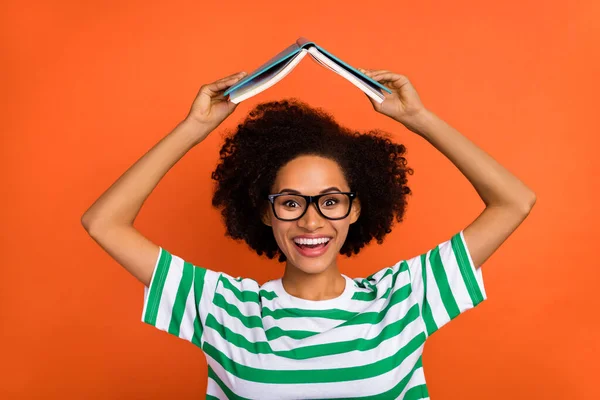 Ritratto di attraente allegra ragazza dai capelli ondulati intelligente tenendo libro sopra la testa imparare isolato su sfondo di colore arancione brillante — Foto Stock