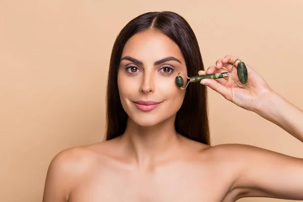 Portrait of attractive brunet girl massaging smooth skin under eyes lifting effect solution isolated over beige pastel color background — Stockfoto