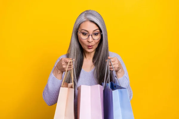 Photo of impressed funny lady pensioner dressed knitted pullover looking open packages isolated yellow color background — стоковое фото