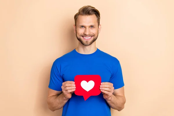 Photo of young cheerful man hold heart icon share repost comment app isolated over beige color background — 스톡 사진