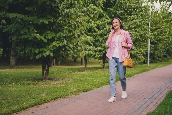Foto de mujer jubilada dulce divertida camisa de rayas vestida hablando dispositivo moderno caminando fuera del paisaje del campo —  Fotos de Stock