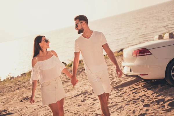 Молодая пара с улыбкой проводит выходные на песчаном пляже возле моря, держа руки в солнцезащитных очках — стоковое фото