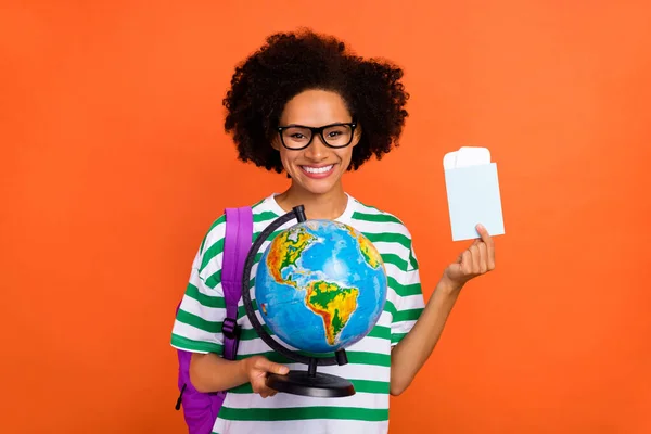 Portret van aantrekkelijke vrolijke golvende harige meisje leerling houden globe pass vliegticket geïsoleerd op fel oranje kleur achtergrond — Stockfoto