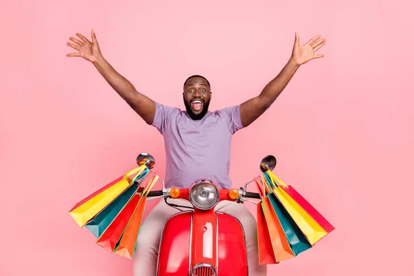 Photo of cheerful impressed guy wear purple t-shirt riding moped carrying shoppers rising arms isolated pink color background — Stock fotografie