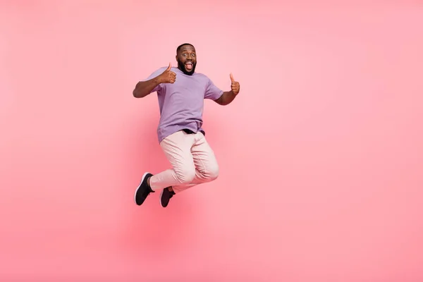 Фото взволнованного симпатичного парня носить фиолетовую футболку прыгая высоко большие пальцы вверх пустое пространство изолированный розовый цвет фона — стоковое фото