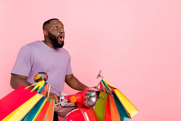 Photo of excited shocked guy dressed violet t-shirt driving moped holding bags empty space isolated pink color background — Stock fotografie