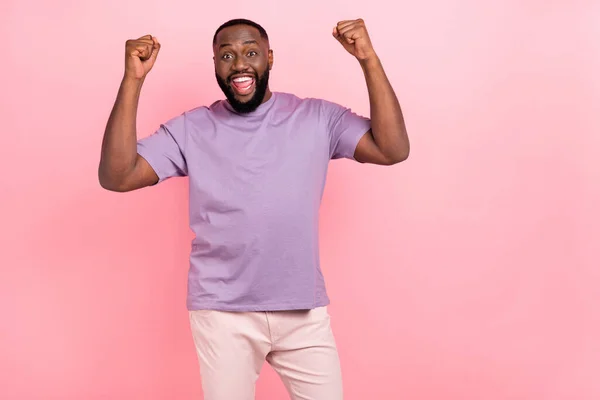 Photo of funny lucky young guy wear purple t-shirt rising fists empty space isolated pink color background — Stockfoto