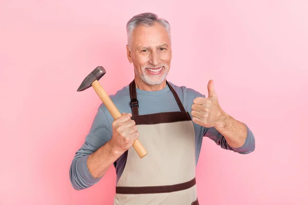 Foto de encantador hombre confiado pensionista vestido gris camisa delantal pulgar hasta martillo aislado color rosa fondo — Foto de Stock